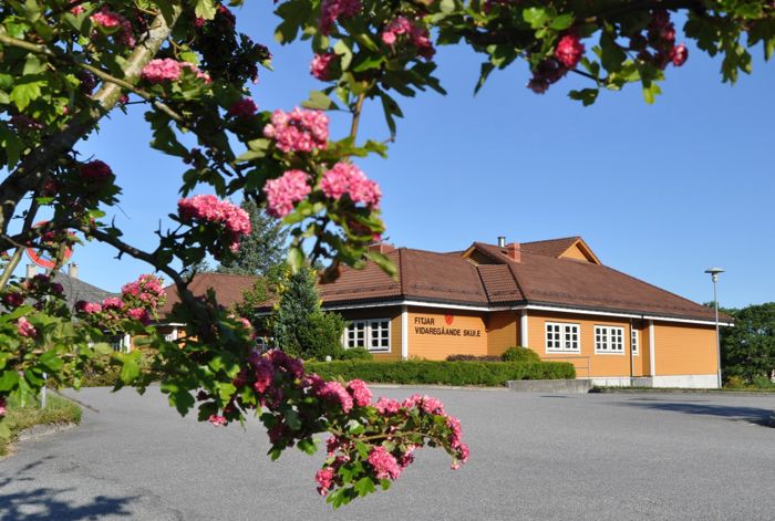 Skulen sett utanifrå. Det er sol og i framgrunnen er det eit tre som blømer.