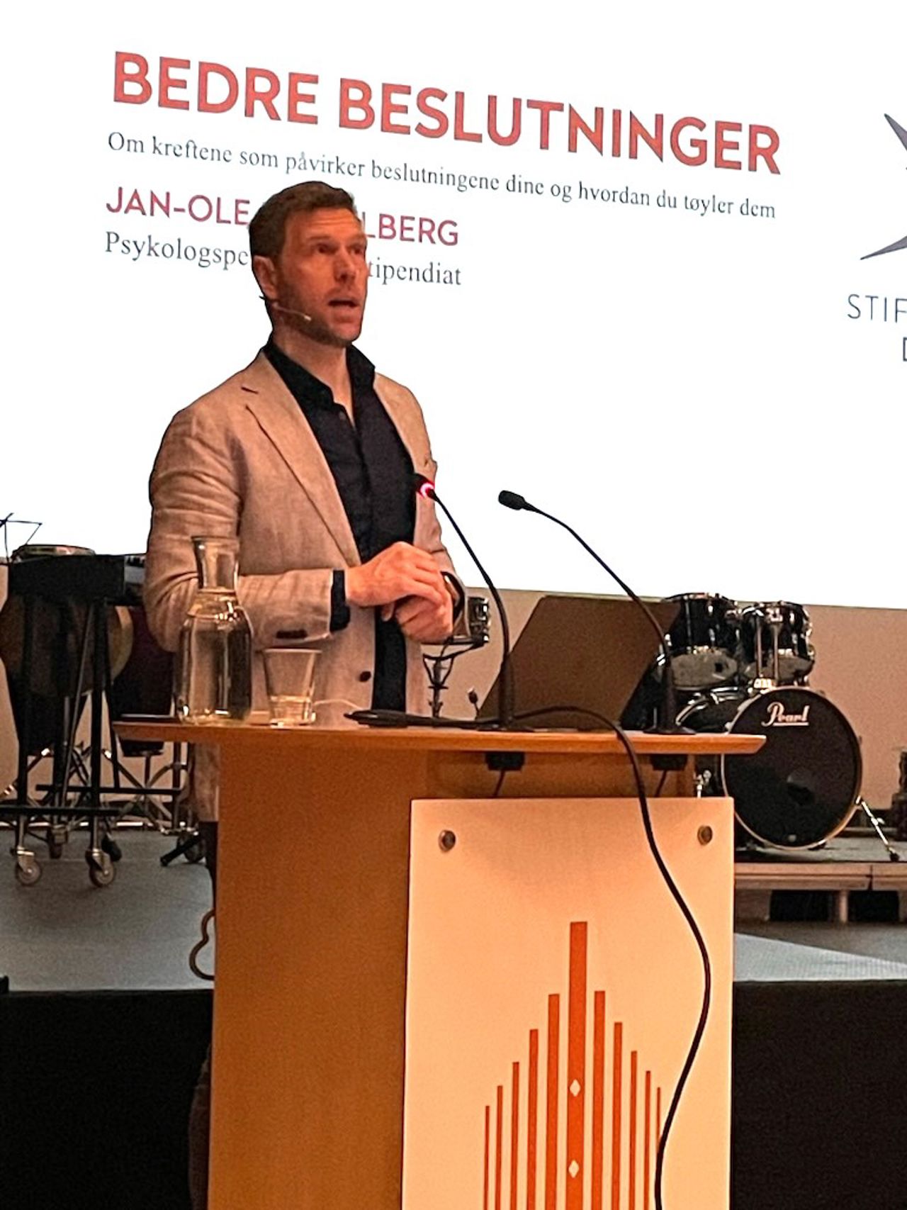 Føredragshaldar Jan-Ove Hesselberg står på ein talerstol og snakker til salen. I bakgrunnen står det skrive "bedre beslutninger."