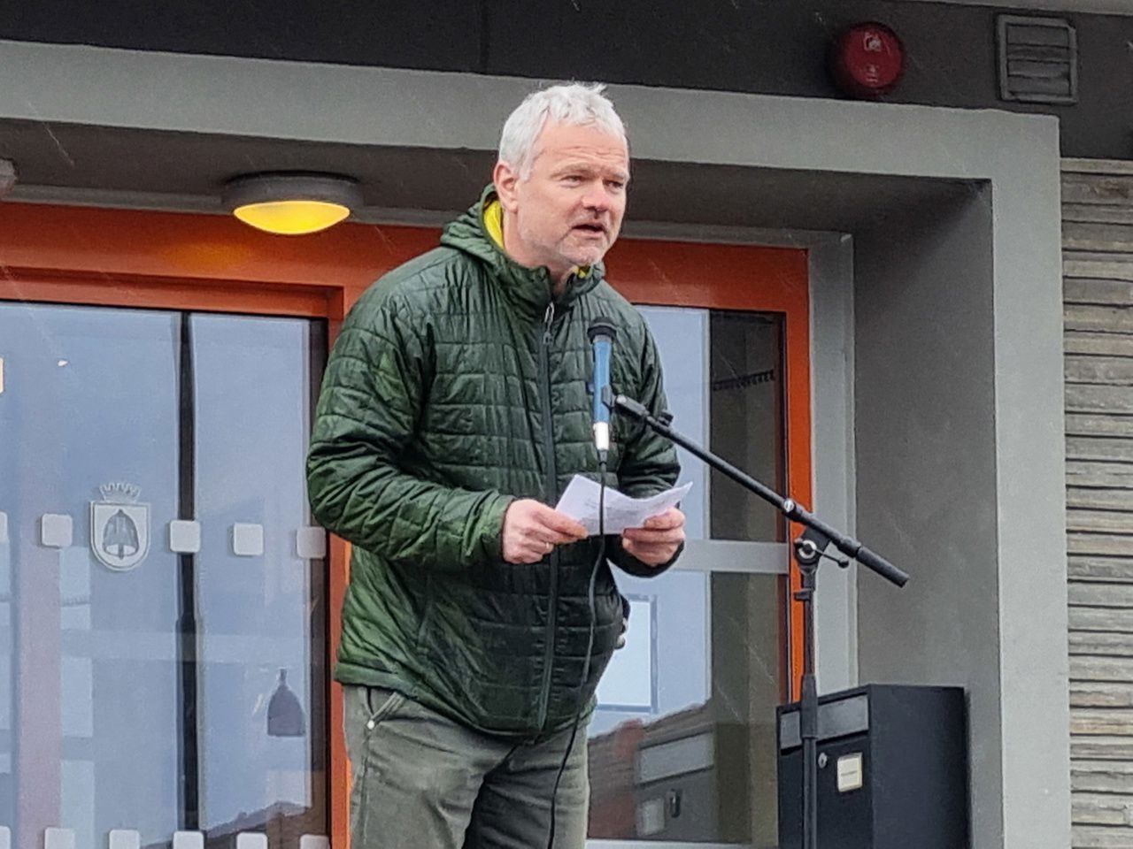 Ein mann i grøn jakke som held ein tale framfor ein mikrofon.