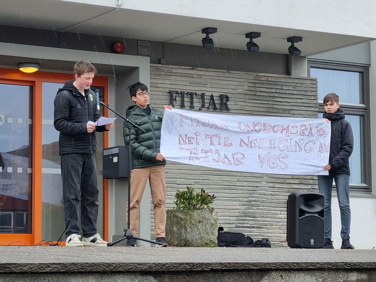 Ein ungdom som held ein tale framfor ein mikrofon. Attmed står det to andre ungdommer og held i eit banner der det står "Fitjar ungdomsråd. Nei til nedlegging av Fitjar vgs"
