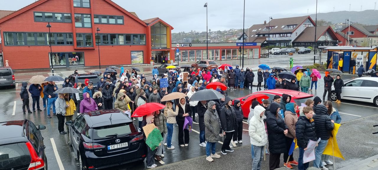 Ei mend av folk med paraplyar og demonstrasjonsplakatar som står på ein parkeringsplass og høyrer på appellar