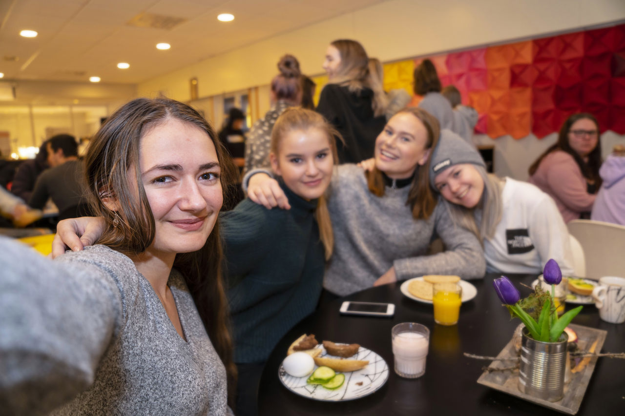Fire jenter som sit i kantina og held rundt kvarandre og smiler, mat står på bordet.